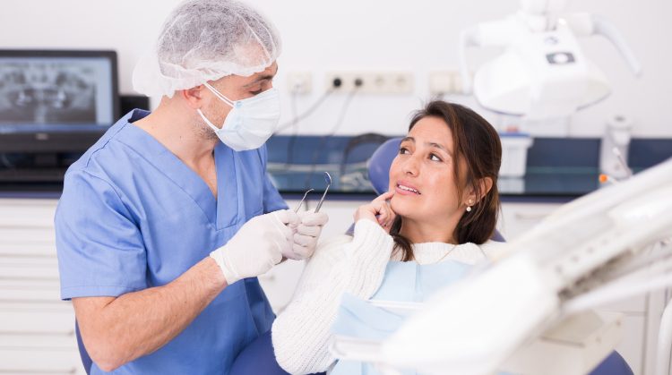 Dentist examining a patient's teeth in a dental office. The patient is seated in a dental chair, smiling, while the dentist uses a dental mirror and explorer for the examination.