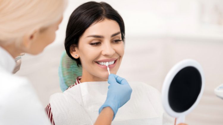 A smiling woman at a dental clinic receiving a professional teeth whitening treatment while holding a mirror, assisted by a dentist in gloves.